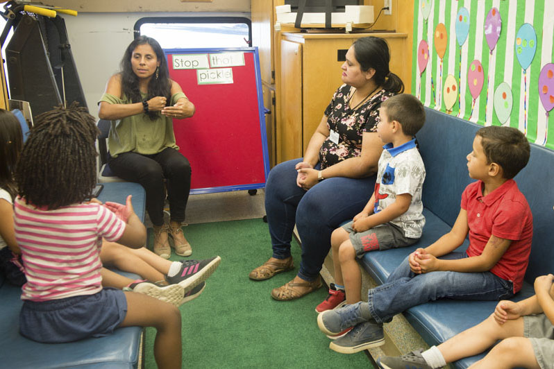 A mixture of children and adults sit on padded benches inside a book mobile listening to a woman tell a story. Photo credit Becky Ruppel.