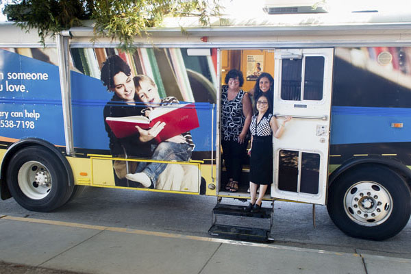 Large trailer book mobile with a picture of a woman reading to a child on her lap on the side. Three women smile from the doorway. Photo credit Becky Ruppel.