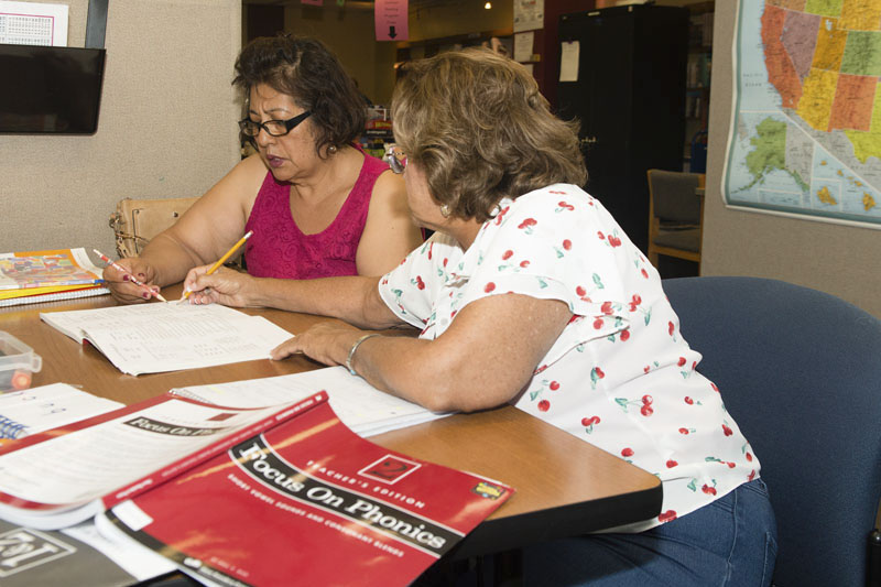 Two women are sitting at a table looking at a book. One woman is pointing at the page with a pencil while the other leans across and writes something. Focus on Phonics book is in the foreground.