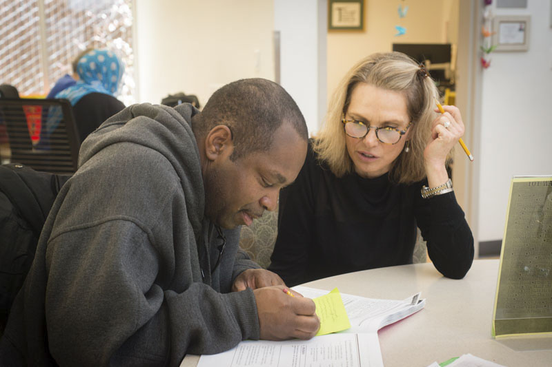 A man sits at a table writing while a woman sits next to him watching and commenting. Photo credit Becky Ruppel.