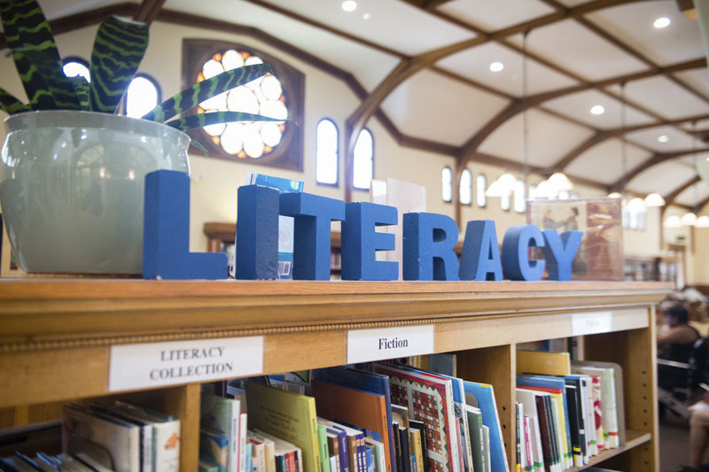 Blue Block sign reading literacy is sitting on top of a book shelf filled with books. Photo Credit Becky Ruppel.