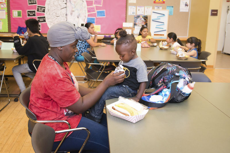 A small child sits on a table next to a hot dog drinking milk from a straw while a woman sitting at the table holds the container for him. Other children and caregivers sit at tables in the background. Photo Credit Becky Ruppel.