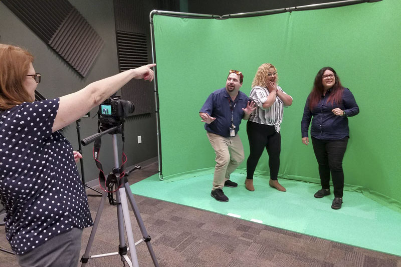 A woman with a video camera on a tri-pod videotaping three adults posing against a green screen. Photo credit the get Involved Project.