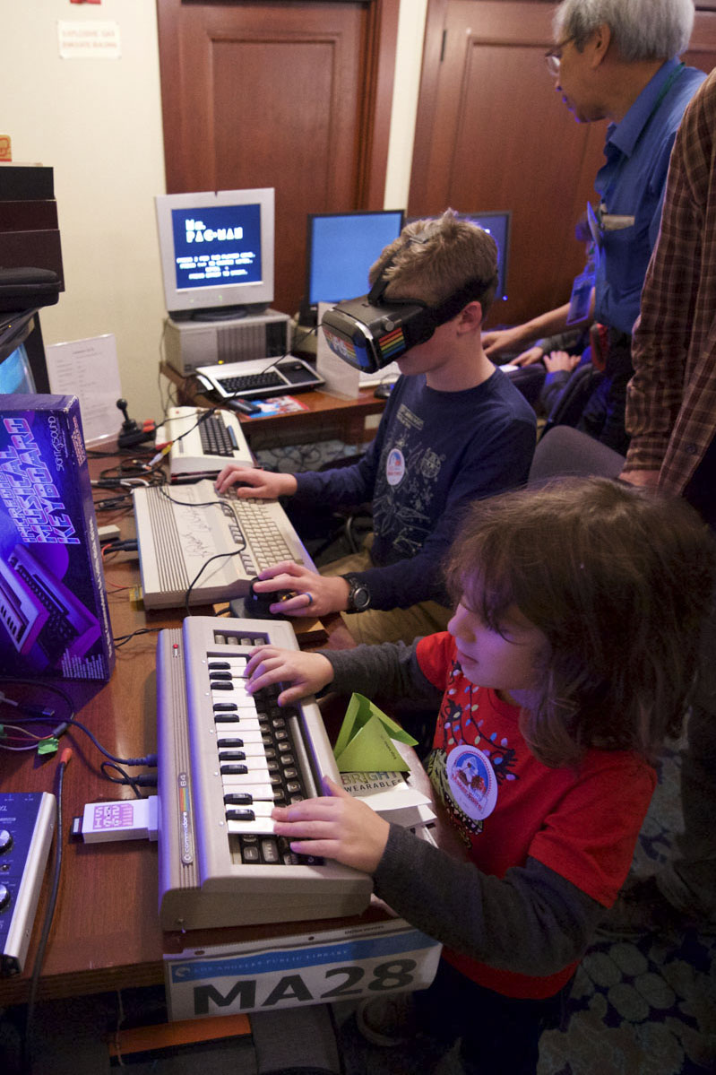 Two kids using technical equipment. The one in the foreground is watching a screen and using a keyboard while the one in the back ground is wearing VR goggles and using a mouse and Keyboard system. Photo courtesy of the Los Angeles Public Library.