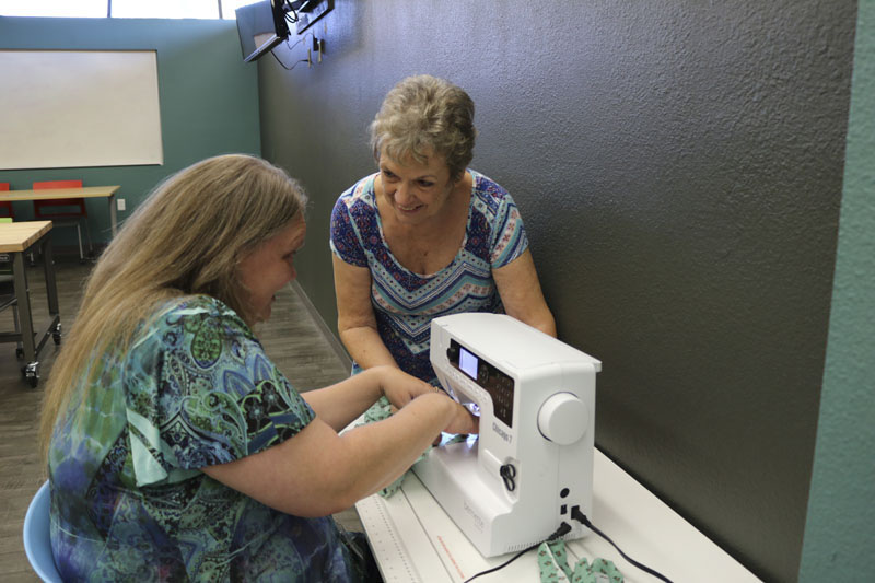 One woman sits at a sewing machine while another woman leans over watching.