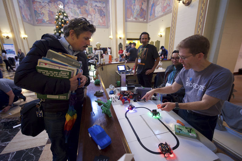 A mom holding an arm full of materials is looking at a display of small robots with glowing lights set-up on a tape track. Another man in an “I Love Robots” shirt is pointing to the robots and talking. Photo Courtesy of the Los Angeles Public Library.