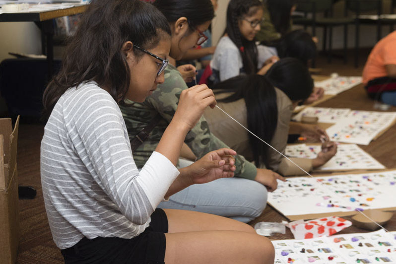 A group of children and young adults sit on the floor using string and other materials to complete worksheets in front of them. Photo Credit Becky Ruppel.