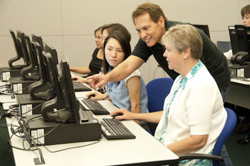 Adults sit at computers set-up in rows in a computer lab setting. A staff member is leaning over a woman’s shoulder and showing her something on the screen. Photo credit the Get Involved Project.