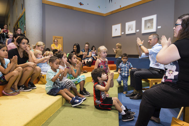 A large group of kids and adults sit on colorful steps facing an in-set stage where a man and woman lead them in hand motions. Photo credit Becky Ruppel.