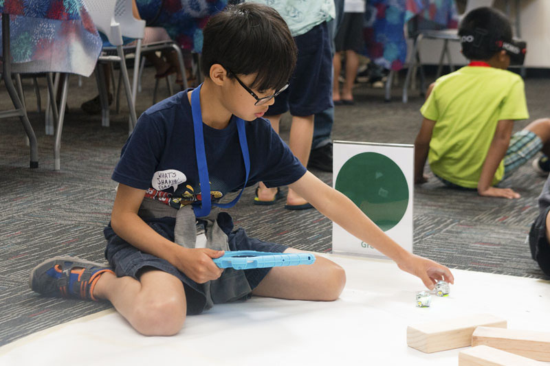 A boy sits on the ground holding a tablet and adjusts a small robot in front of him. Photo credit Becky Ruppel.