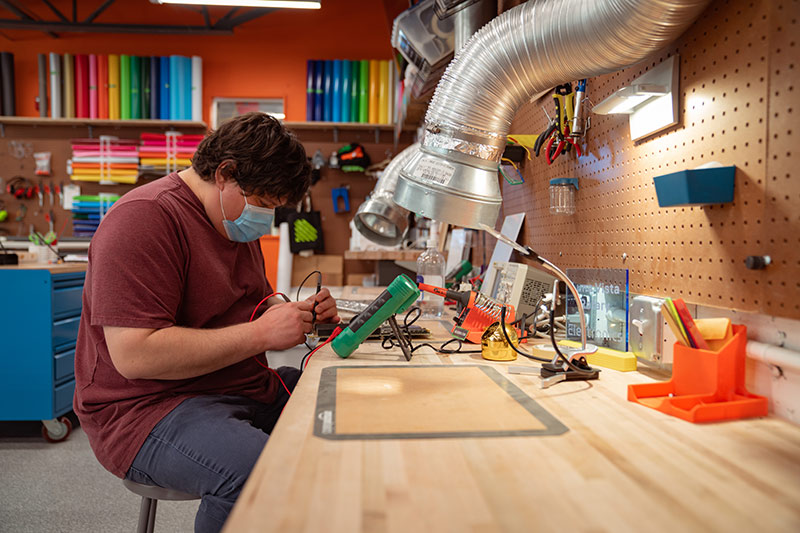 Young adult man sitting at a bench working with electronics. Photo credit Christian Koszka.