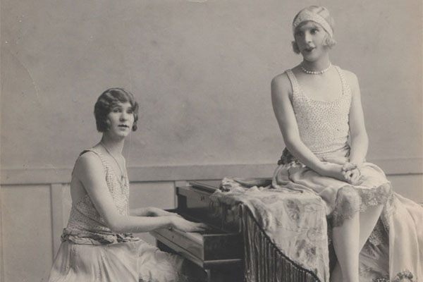 Female Impersonators in 1920s attire, one playing piano, one sitting atop.