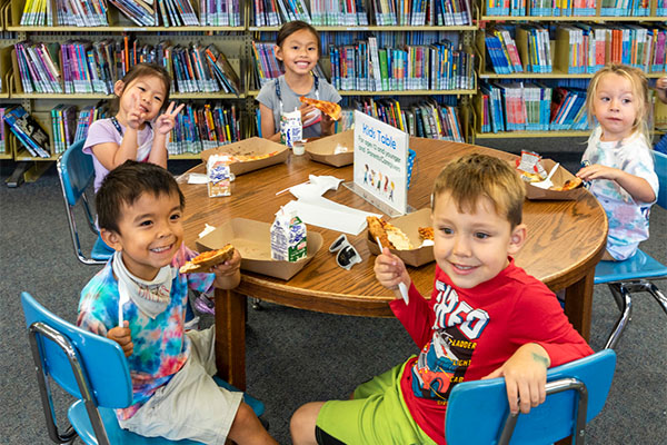 Five kids in a library eating food at a small table.