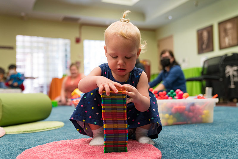 Young child stacking magnetic tiles on the floor while two adults observe from afar.