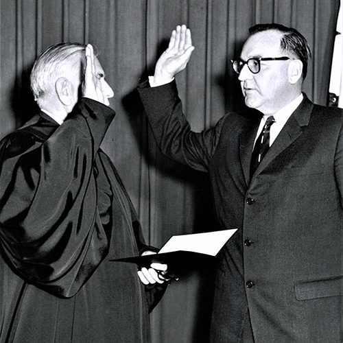 Governor Edmund G. Brown pictured taking the oath of office from California Supreme Court Justice Phil Gibson at the State Capitol during the 1959 Inauguration. Both men are holding up their right hands and are standing in front of a draped wall.